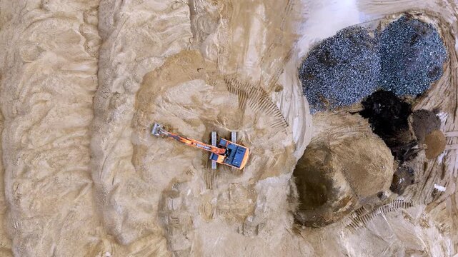 Aerial view shows sandy terrain with a backhoe digging a circular pit. Loose soil forms a deep trench as the machine works. Rocky outcrops surround the excavation site