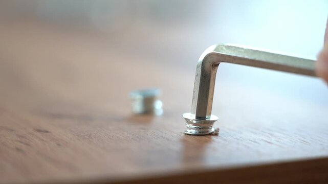 Two metal pins lie on a wooden surface. Shallow focus emphasizes the pins. Soft sunlight highlights the scene gently. Metal tool touches one pin. Hand guides it into the pin. The action is precise