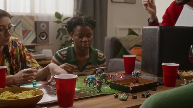 Close-up of friends playing tabletop RPG, focused players studying game board while game master behind screen gesturing with pen and running story in cozy living room