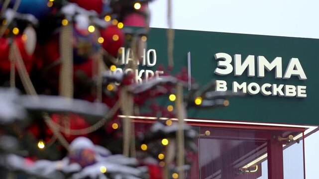 Moscow, December 30 2025, Green sign displays 'Winter in Moscow' in Cyrillic letters. Festive lights twinkle near snow-covered branches. Snow blankets the scene softly. This captures Moscow's winter