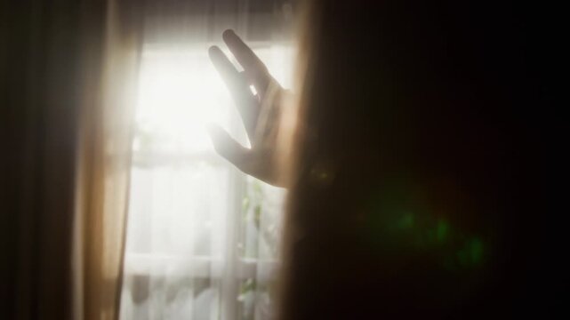 Dreamy over shoulder shot of unrecognizable young girl waving hand against sunlight coming through lace curtains on window and daydreaming in cozy home setting, copy space