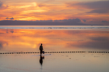 Fototapeta premium A lone figure, stands in an endless mirror of sunrise, on the beach in Bali, Indonesia, with the burnt orange sky reflecting in the ocean to create a magical landscape