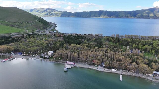 Aerial drone view of mountain lake shoreline with forest and winding road under bright cloudy sky 4K.