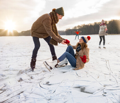 Man helping ice skating woman up on icy surface of frozen lake
