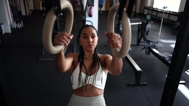 Focused young woman with braided hair doing strength exercises on gymnastic rings. Intense workout session building upper body and core strength, demonstrating dedication and fitness