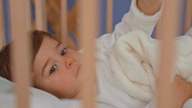 Boy in wooden crib lies on back reaching hand to soft white blanket while gazing upward inside a building; calm comfort.