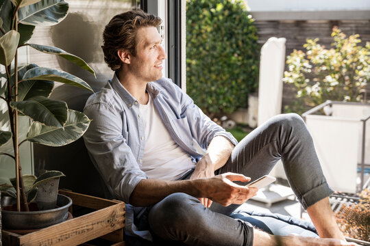 Mid adult man holding smart phone looking away while sitting at home