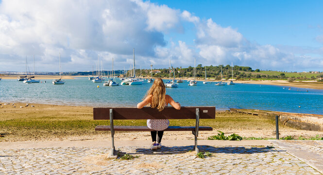 Woman sitting on a wooden bench looking at the river estuary and boats in Alvor, Algarve, Portugal