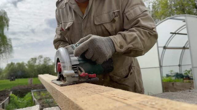 Carpenter cutting a wooden board with a circular saw outdoors. Close-up of a worker in a protective jacket and gloves using an electric circular saw to cut a timber plank. 