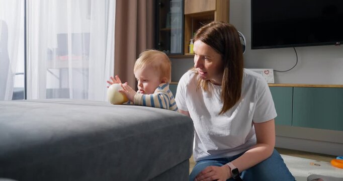 A mother interacts and plays with her curious toddler who is exploring and holding a round object in a cozy living room setting, showcasing early childhood development and bonding time