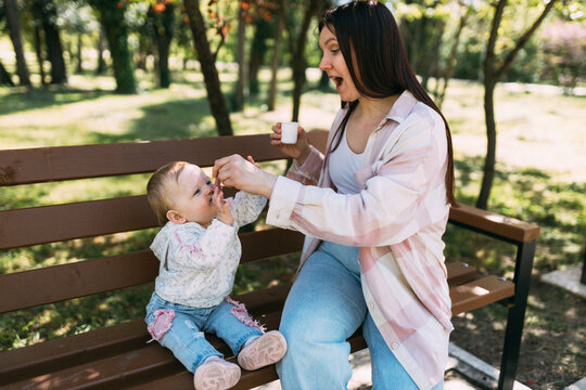 Mother feeding baby daughter on park bench during outdoor walk