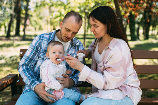 Parents feeding baby daughter together on park bench outdoors