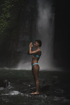 Serene woman in bikini with prayer hands by misty waterfall, Bali