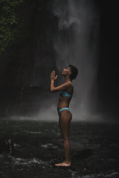 Woman in bikini in prayer pose looking up at waterfall, Sekumpul, Bali