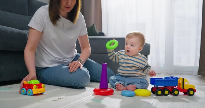 A mother playing and engaging with her baby using colorful stacking rings and a toy truck on a carpeted floor in a bright, cozy living room setting