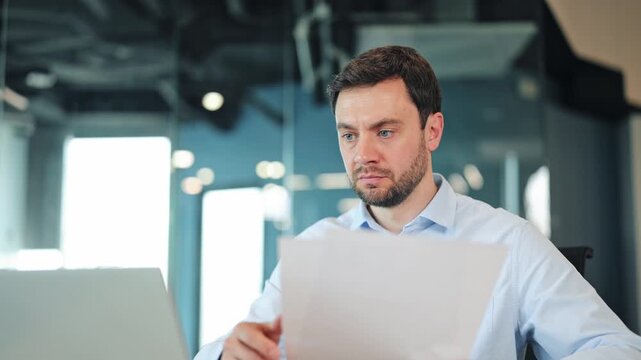 Focused businessman or IT specialist checking corporate documents at desk. Happy manager, worker or freelance designer holding paper report and smiling at camera in contemporary coworking workspace.