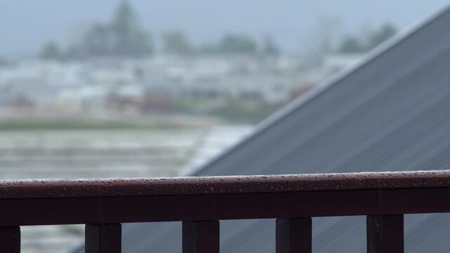 Heavy rain hitting a wet brown wooden balcony railing covered in water droplets. A dark metal roof and a blurred town landscape sit in the background. Static eye level view.