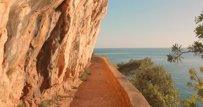 Narrow dirt trail hugging dramatic ochre cliffs above the Mediterranean Sea. Lush coastal vegetation and turquoise water visible.