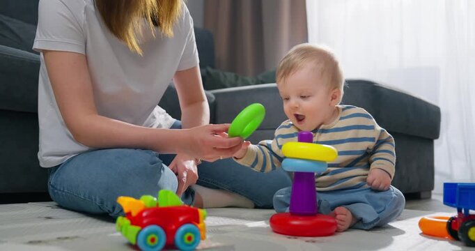 A baby playing with colorful stacking rings while sitting on the floor next to an adult, engaging in early developmental play with toys alongside a red and yellow toy car in a cozy living room setting