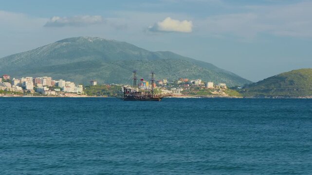 Black sailboat cruising coastal mountain, static tripod shot. Traditional sailing vessel full of people navigating on sea water in front of city town houses.