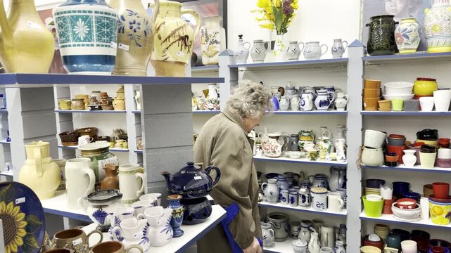 An elderly woman is looking around a thrift store; she is in the glass and china section, panning shot