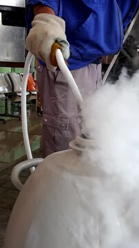 A worker wearing protective gloves and blue jacket handles a hose releasing vapor from a large liquid nitrogen container indoors.