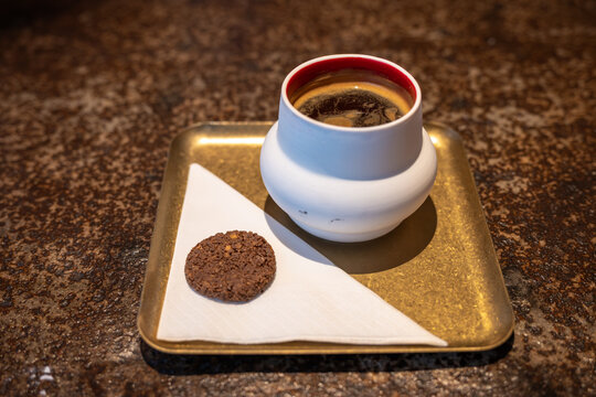 A steaming ceramic cup of black coffee served on a brass tray with a chocolate cookie on a folded napkin, creating a cozy, rustic cafe mood and simple breakfast moment.