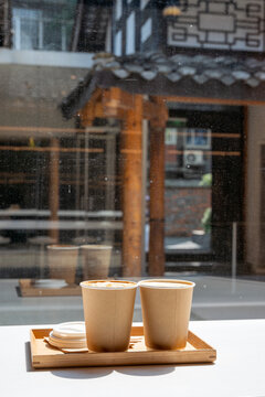 Two takeaway coffee cups with latte art sit on a wooden tray by a sunlit cafe window, evoking cozy moments, casual conversation, and urban slow mornings.