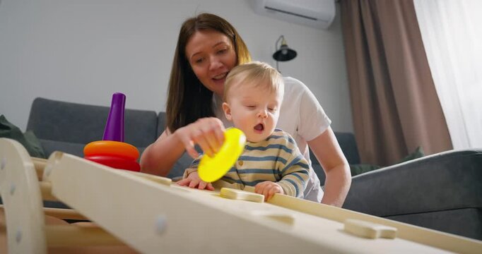 Mother engaging with her toddler child in a developmental play activity using colorful ring stacking toys and wooden educational objects in a cozy living room setting with natural light