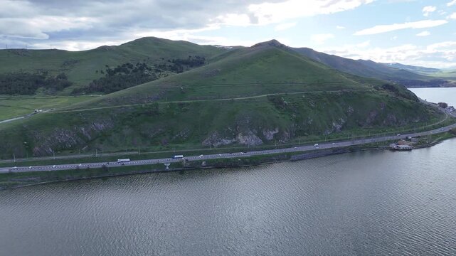 4K Drone Aerial View of Scenic Mountain Road Along Large Blue Lake With Green Hills and Natural Landscape.