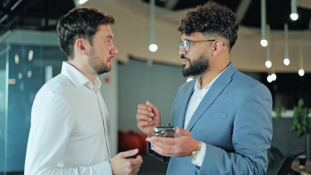 Two young male professionals looking at mobile phone screen in modern open space office. Company colleagues or startup founders analyzing internet statistics or online app.