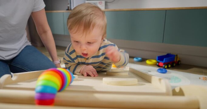 Curious toddler playing with colorful toys and wooden puzzle pieces on the floor while an adult watches closely in a modern home environment featuring cabinets and scattered play objects