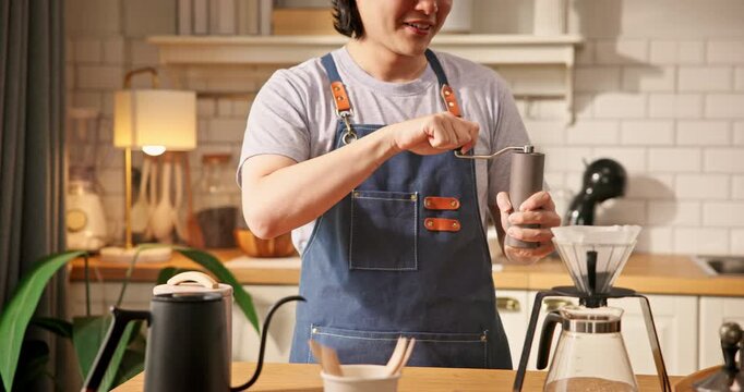Young Asian home barista in apron using manual hand grinder to grind coffee beans. Smiling gen-z male preparing fresh coffee in kitchen. Slow life, specialty coffee hobby, and morning routine concept.