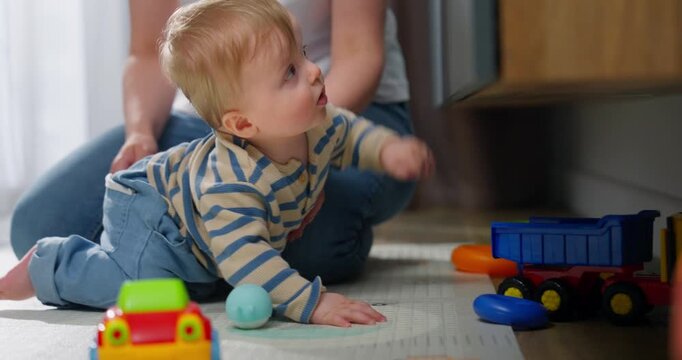 A young toddler playing and crawling on the floor with colorful toy cars while being closely supervised by an adult sitting nearby in a cozy indoor setting