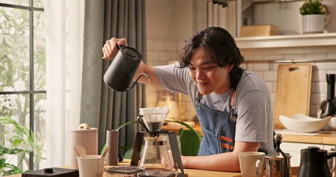 Young Asian man pouring hot water from kettle into glass dripper with paper filter. Home barista brewing specialty pour over coffee in morning kitchen. Hand drip coffee brewing process hobby lifestyle