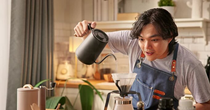 Young Asian man pouring hot water from kettle into glass dripper with paper filter. Home barista brewing specialty pour over coffee in morning kitchen. Hand drip coffee brewing process hobby lifestyle