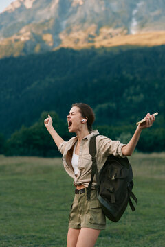 A young woman in casual clothes with a backpack stands outdoors in a green field with mountains behind her, shouting joyfully with arms raised. Lifestyle and freedom concept.