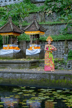 Young Balinese legong dancer in gold headdress and green batik costume standing on mossy stone steps at temple pond
