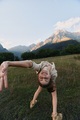 Obraz premium A smiling child performs a backbend in a meadow with mountains in the background, capturing carefree outdoor play, movement and balance in a bright nature scene.