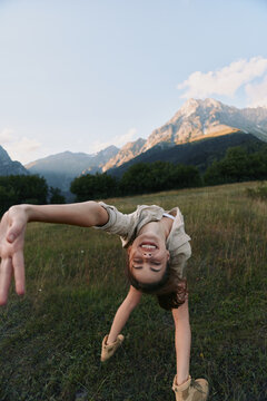 A smiling child performs a backbend in a meadow with mountains in the background, capturing carefree outdoor play, movement and balance in a bright nature scene.