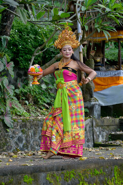 Balinese female dancer in elaborate golden crown and colorful sarong posing beside lotus pond with temple pavilions