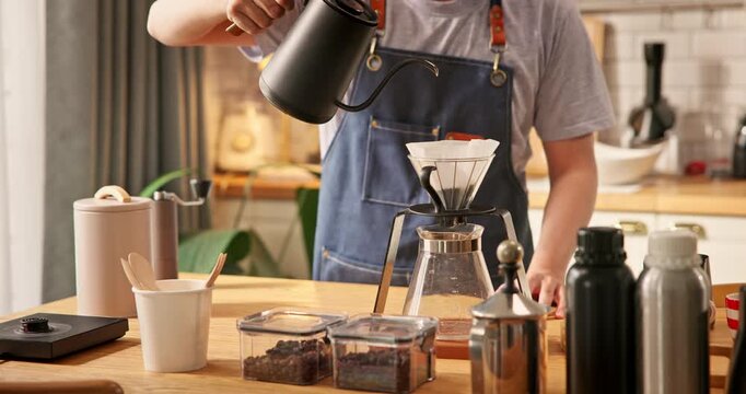Young Asian man pouring hot water from kettle into glass dripper with paper filter. Home barista brewing specialty pour over coffee in morning kitchen. Hand drip coffee brewing process hobby lifestyle