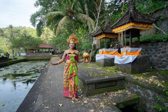 Full body shot of Balinese legong dancer in vibrant traditional costume at stone temple walkway beside lotus water garden