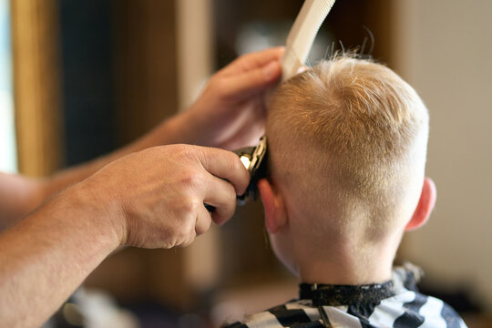 Coiffeur coupant les cheveux d'un petit gar&ccedil;on blond avec une tondeuse &eacute;lectrique et un peigne dans un salon de coiffure ou &agrave; la maison. Gros plan sur les mains du professionnel en action