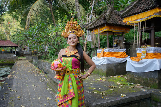 Elegant Balinese dancer in gold headdress and green pink dance costume standing at ornate temple with lotus pond