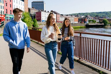 Fototapeta premium Teenagers walking along waterfront with coffee in sunny city
