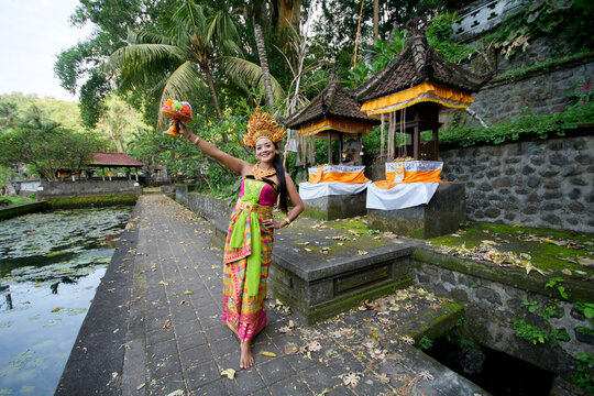 Balinese legong dancer in full golden headdress and colorful costume posing on stone pathway at lotus pond temple