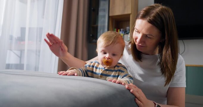 A mother lovingly watches her young child with a pacifier as the baby explores and interacts with a soft surface in a cozy home setting filled with natural light and warmth