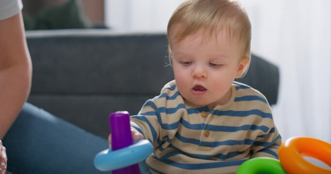 Curious toddler in a striped shirt playing and exploring colorful toys on the floor with adult nearby in a cozy indoor setting, focused on stacking and sensory play activities