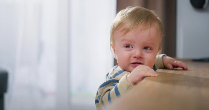 A curious toddler with blonde hair in a striped shirt leaning on a wooden surface in a cozy indoor setting, showing expressions of curiosity and slight distress while being supported by an adult.